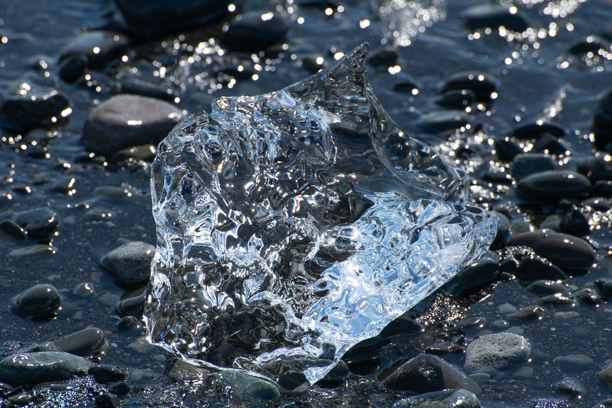 A transparent piece of ice sits on a shore of wet, dark rounded pebbles, with bright sunlight glinting off the water and stones.