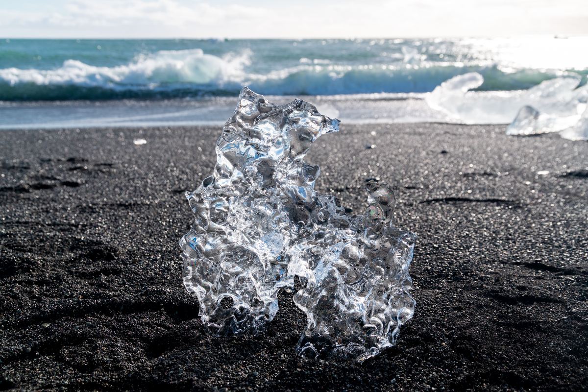 A translucent chunk of ice with a rippled surface sits on dark volcanic sand, with turquoise waves and more ice pieces blurred in the background.