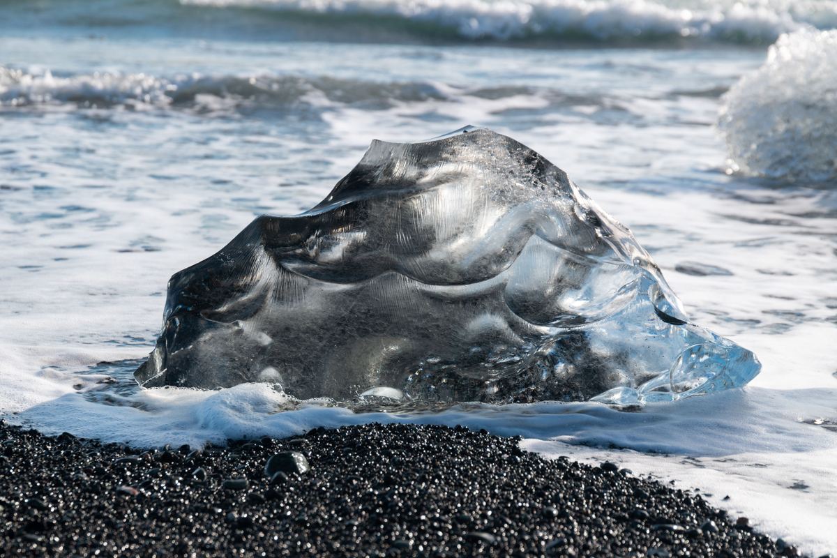 A clear, sculpted piece of ice sits on dark volcanic sand while foamy waves wash around it.
