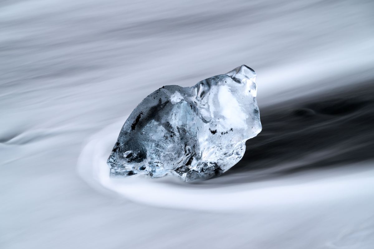 A translucent chunk of ice rests on dark sand as smooth, milky streaks of water swirl around it.