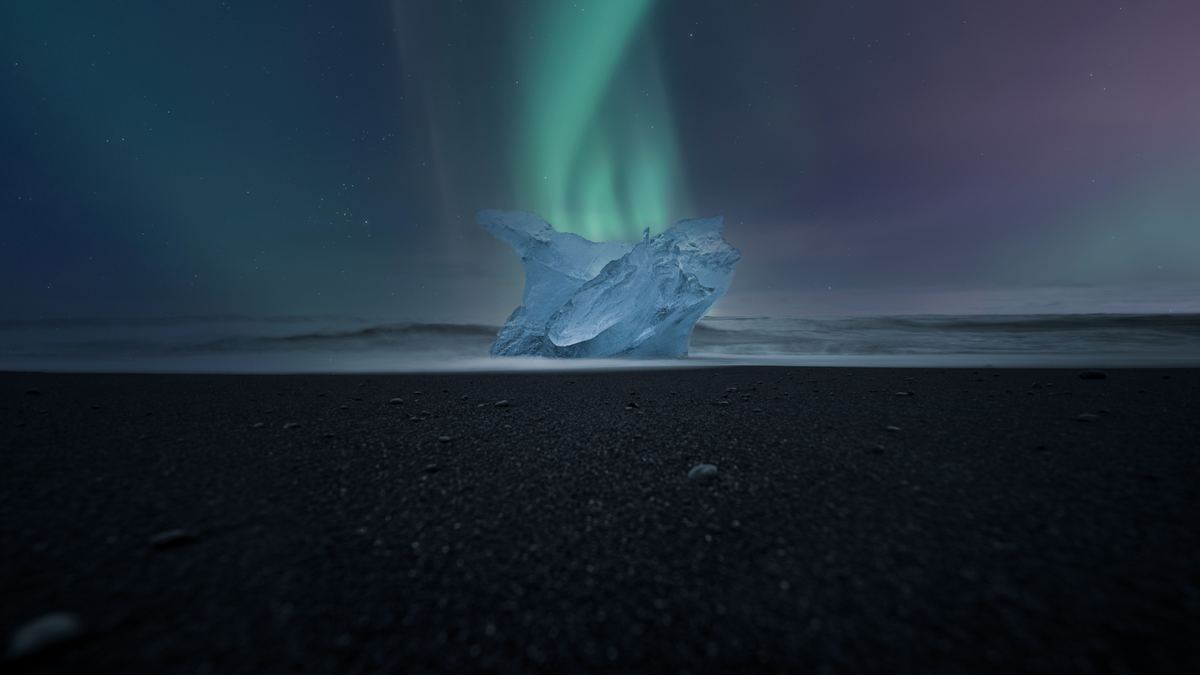 A translucent blue block of ice sits on dark volcanic sand beneath a night sky with a soft green aurora above the sea.