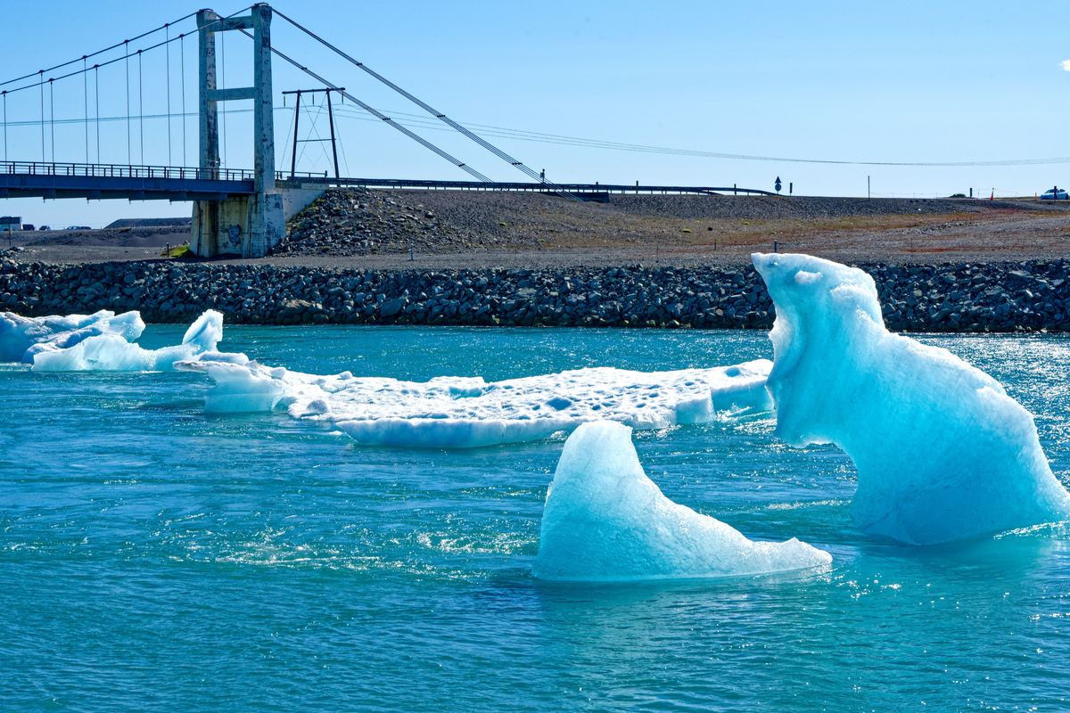 Bright blue icebergs float on turquoise water in front of a rocky shore and a suspension bridge under a clear blue sky.