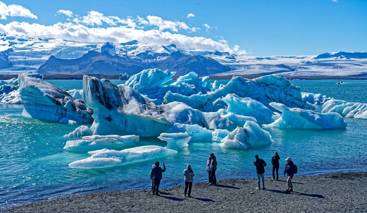 Visitors stand on a dark gravel shore looking at bright blue and white icebergs floating in a turquoise lagoon, with snowy mountains and clouds behind.